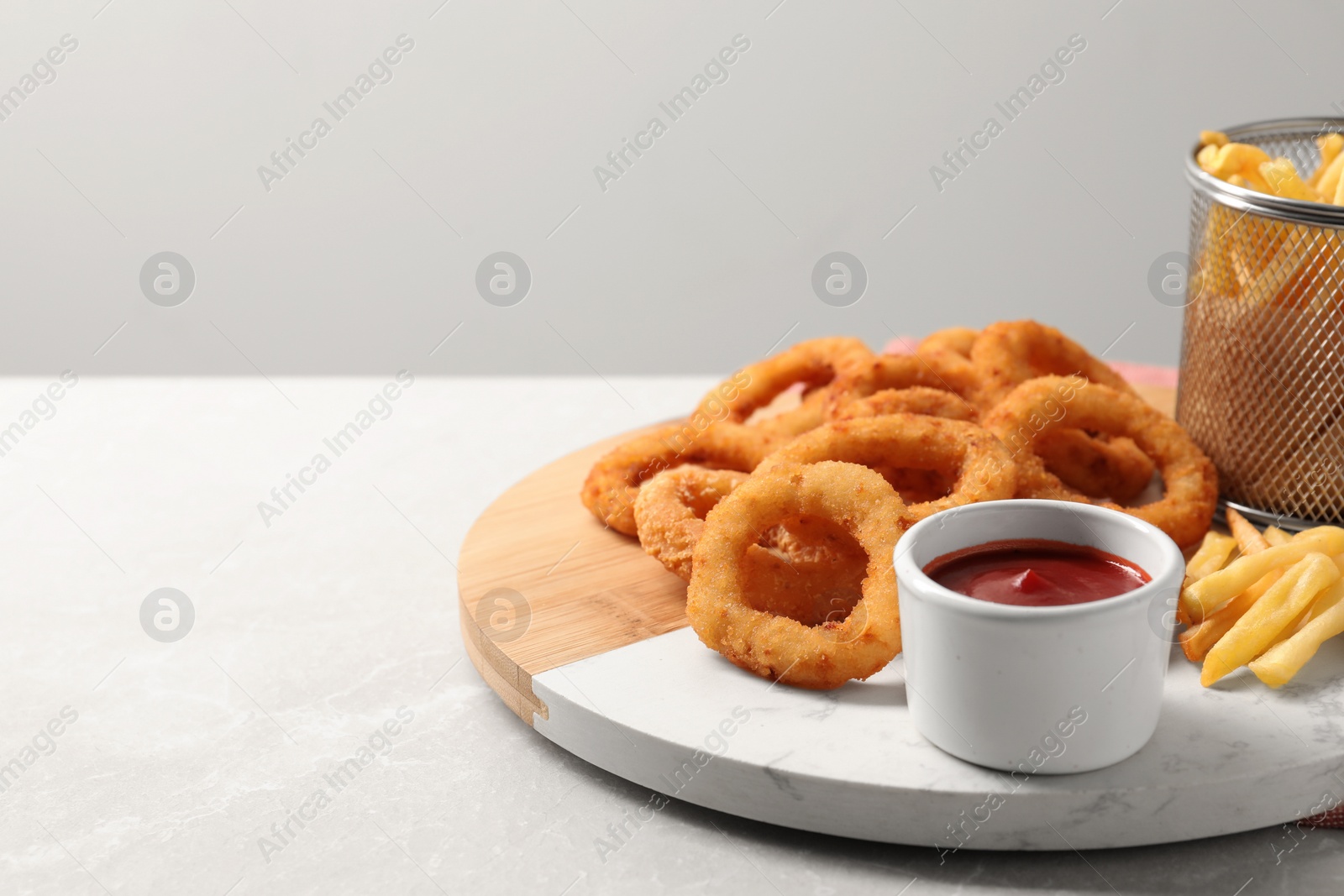 Photo of Board with tasty ketchup, fries and onion rings on light grey table, space for text