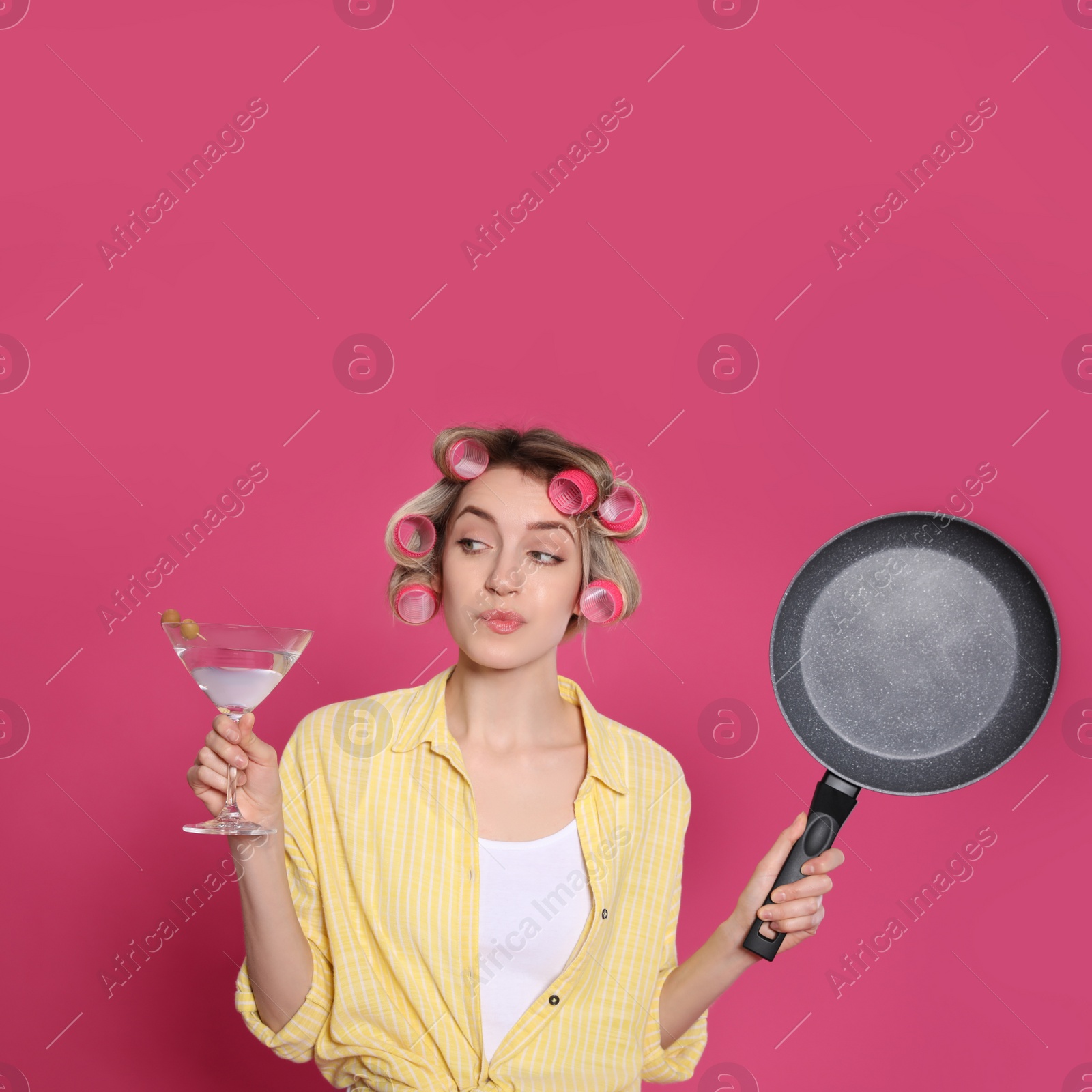 Photo of Young housewife with frying pan and glass of martini on pink background