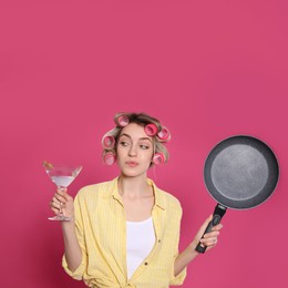 Photo of Young housewife with frying pan and glass of martini on pink background