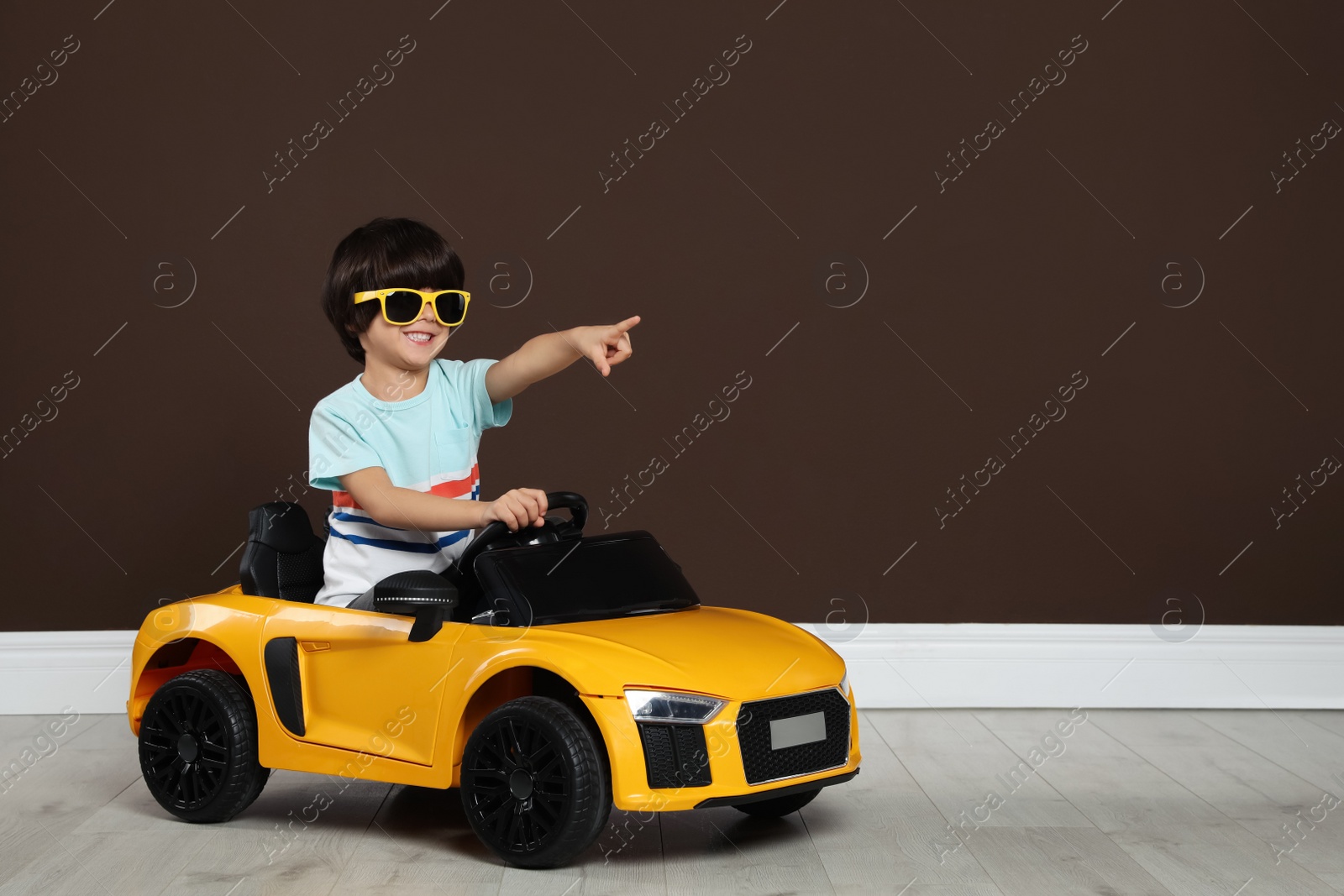 Photo of Cute little boy driving children's electric toy car near brown wall indoors. Space for text