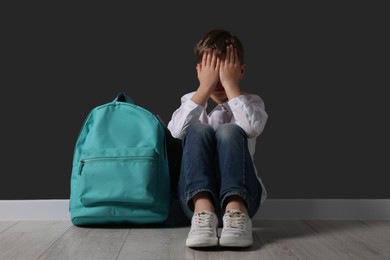 Upset boy with backpack sitting on floor near black wall. School bullying