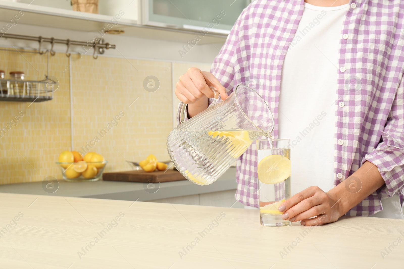 Photo of Young woman pouring lemon water into glass from jug in kitchen, closeup. Space for text