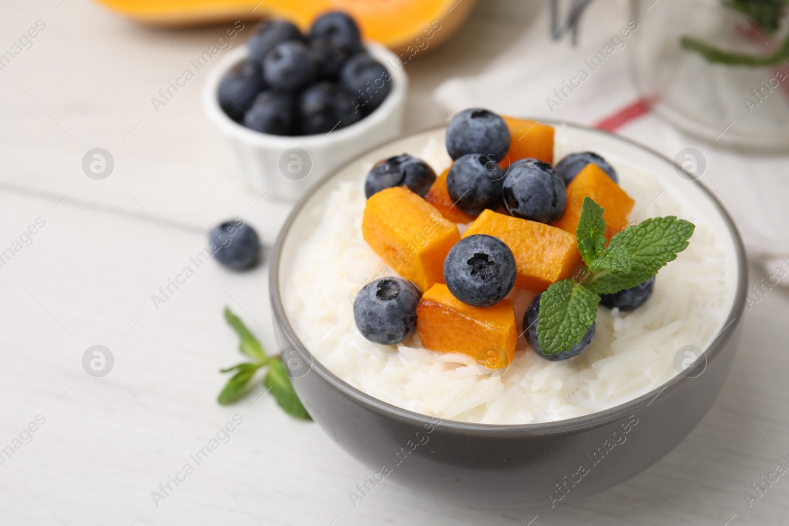 Photo of Bowl of delicious rice porridge with blueberries, pumpkin and mint on white table, closeup