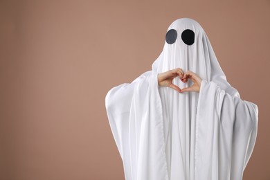 Photo of Cute ghost. Woman covered with white sheet making heart with hands on dark beige background, space for text