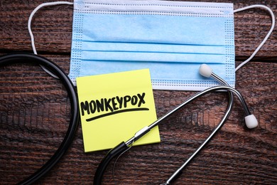 Photo of Note with word Monkeypox, mask and stethoscope on wooden table, flat lay