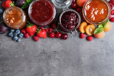 Photo of Jars with different jams and fresh fruits on grey table, flat lay. Space for text