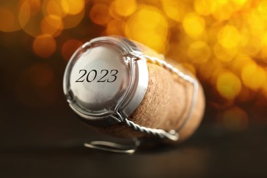 Cork of sparkling wine and muselet cap with engraving 2023 on black table against blurred festive lights, closeup. Bokeh effect