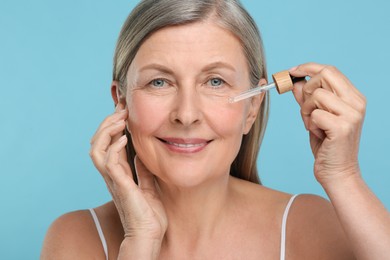 Photo of Senior woman applying cosmetic product on her aging skin against light blue background. Rejuvenation treatment