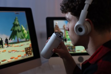Young man with energy drink playing video game at wooden desk indoors, closeup