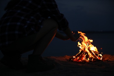 Photo of Woman warming hands near burning firewood on beach at night, closeup