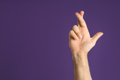 Photo of Man with crossed fingers and space for text on purple background, closeup. Superstition concept