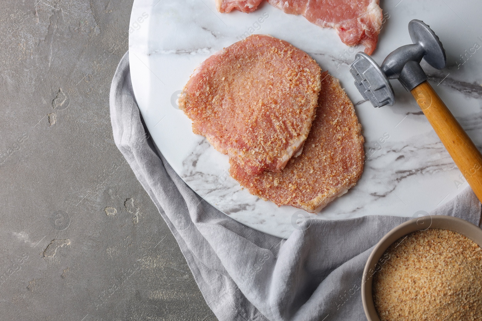 Photo of Cooking schnitzel. Raw pork chops in bread crumbs and meat mallet on grey table, top view with space for text