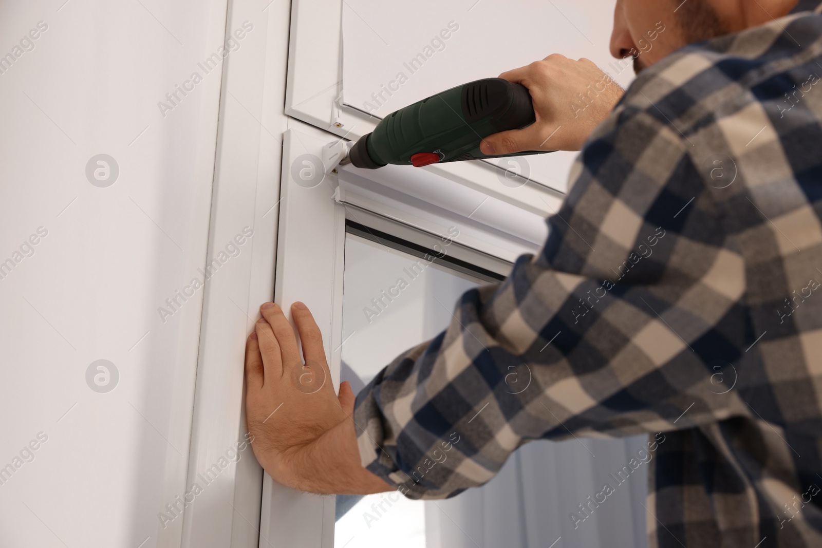 Photo of Man with drill installing roller window blind indoors, closeup