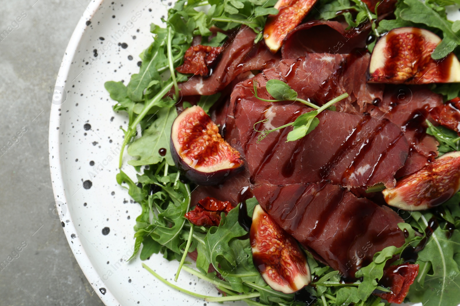 Photo of Plate of tasty bresaola salad with figs, sun-dried tomatoes and balsamic vinegar on grey table, top view