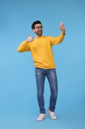 Photo of Smiling man taking selfie with smartphone and showing thumbs up on light blue background