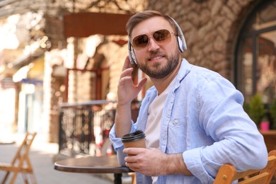 Photo of Happy young man with coffee and headphones listening to music in outdoor cafe