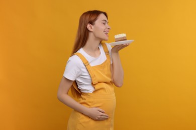 Pregnant young woman eating piece of tasty cake on orange background