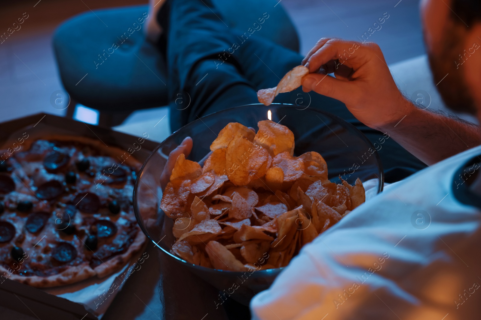 Photo of Man eating chips and pizza while watching TV on sofa at night, closeup. Bad habit