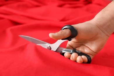 Photo of Professional tailor cutting red fabric with scissors in workshop, closeup