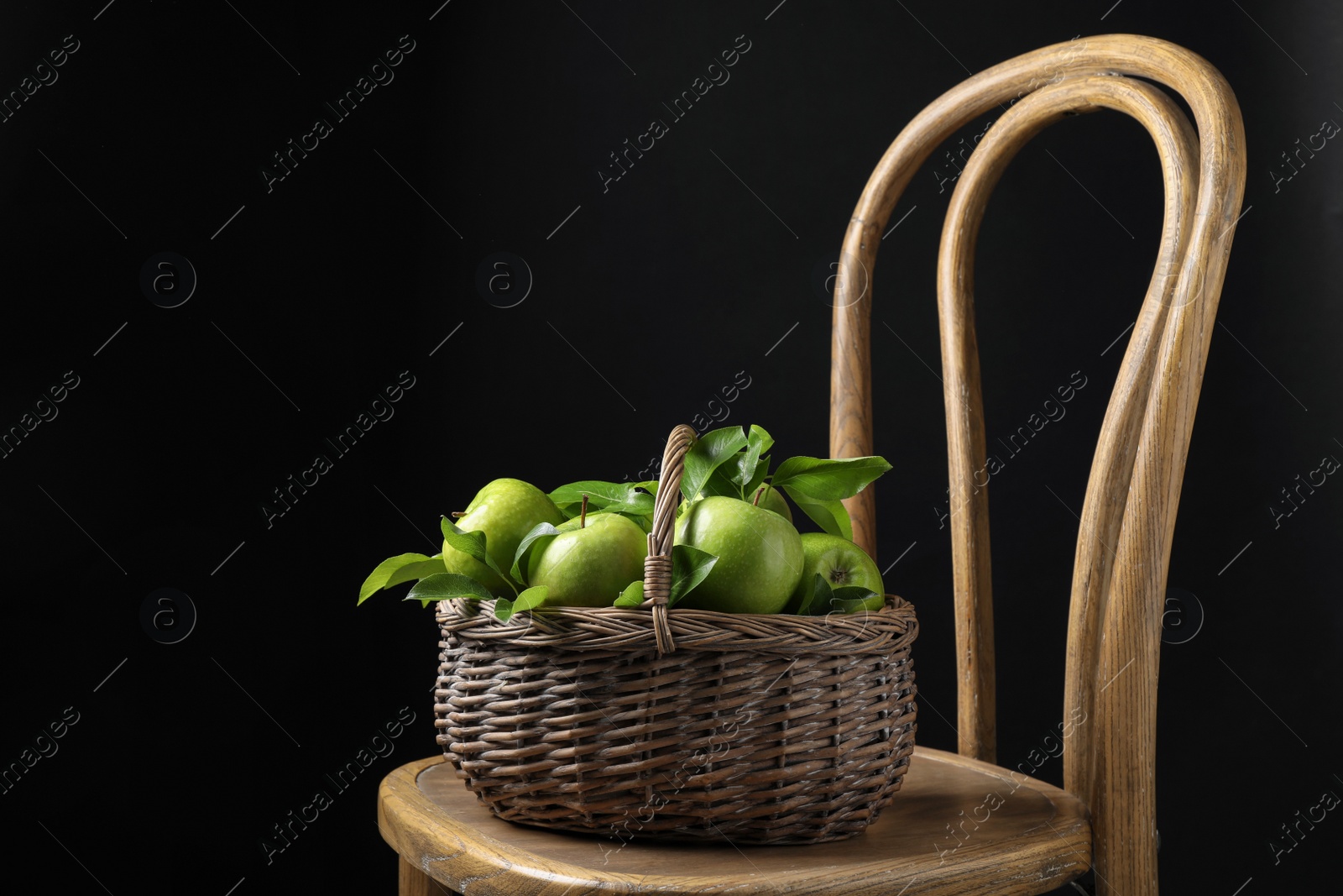 Photo of Fresh ripe green apples and leaves in wicker basket on wooden chair against black background