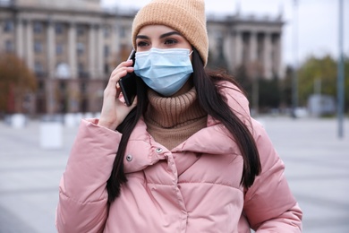 Photo of Young woman in medical face mask talking on phone while walking outdoors. Personal protection during COVID-19 pandemic