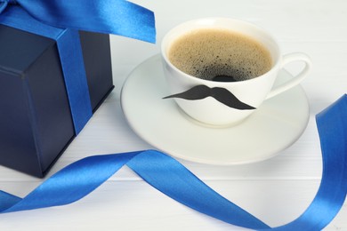Photo of Happy Father's Day. Cup with moustache and gift box on white wooden table, closeup