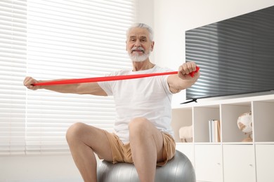 Senior man doing exercise with elastic resistance band on fitness ball at home