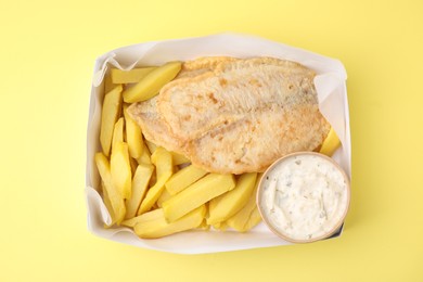 Photo of Delicious fish and chips with tasty sauce in paper box on yellow table, top view
