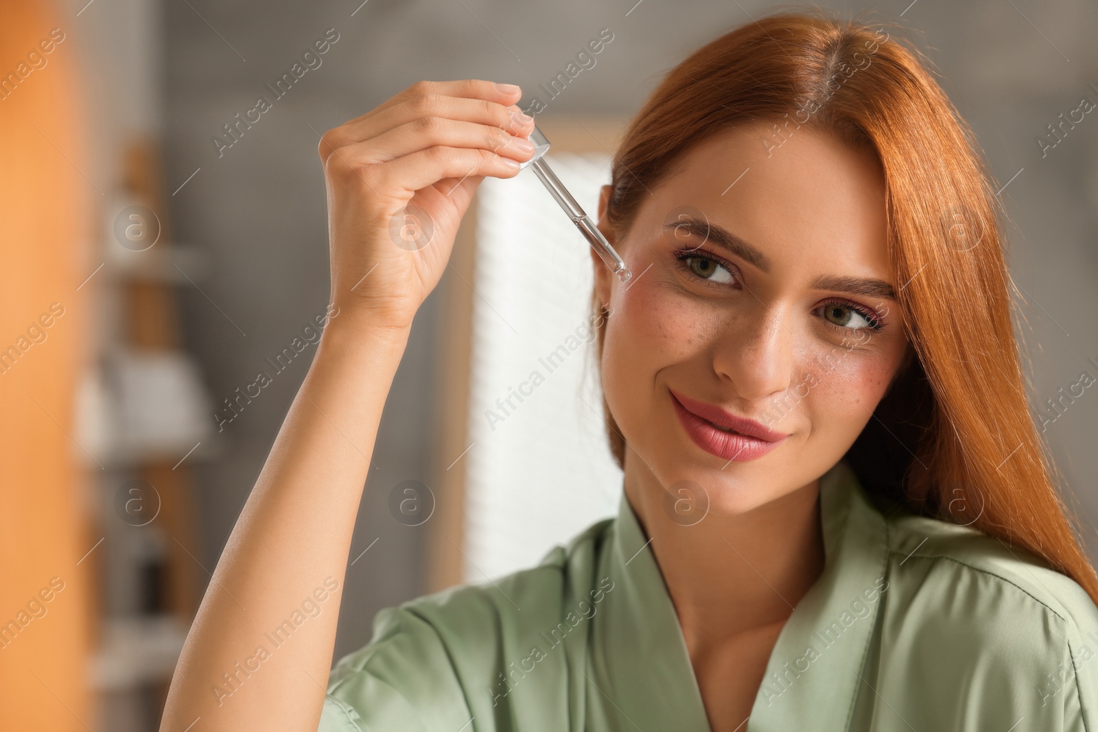 Photo of Beautiful young woman applying cosmetic serum onto her face near mirror in bathroom