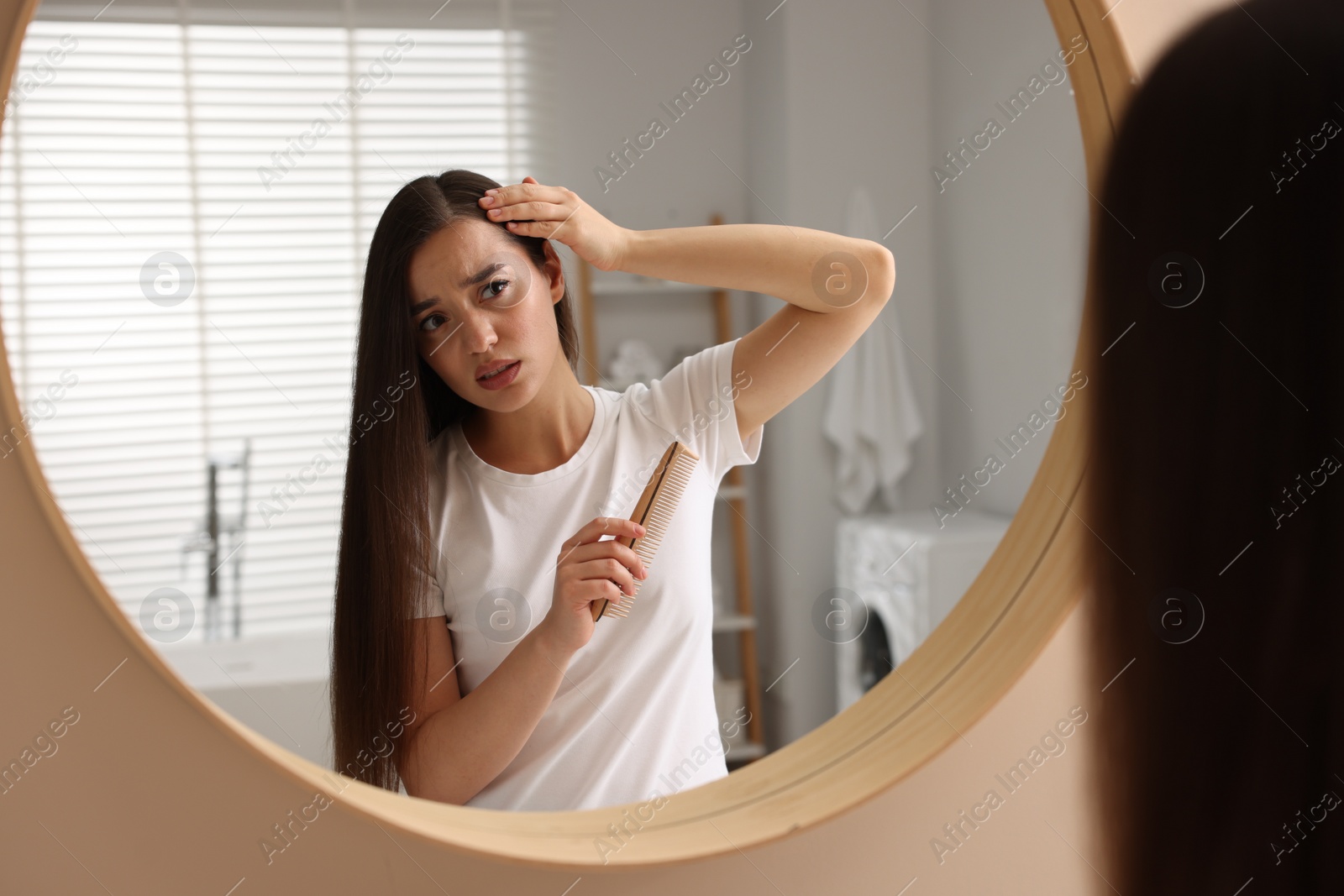 Photo of Emotional woman with comb examining her hair and scalp near mirror in bathroom. Dandruff problem