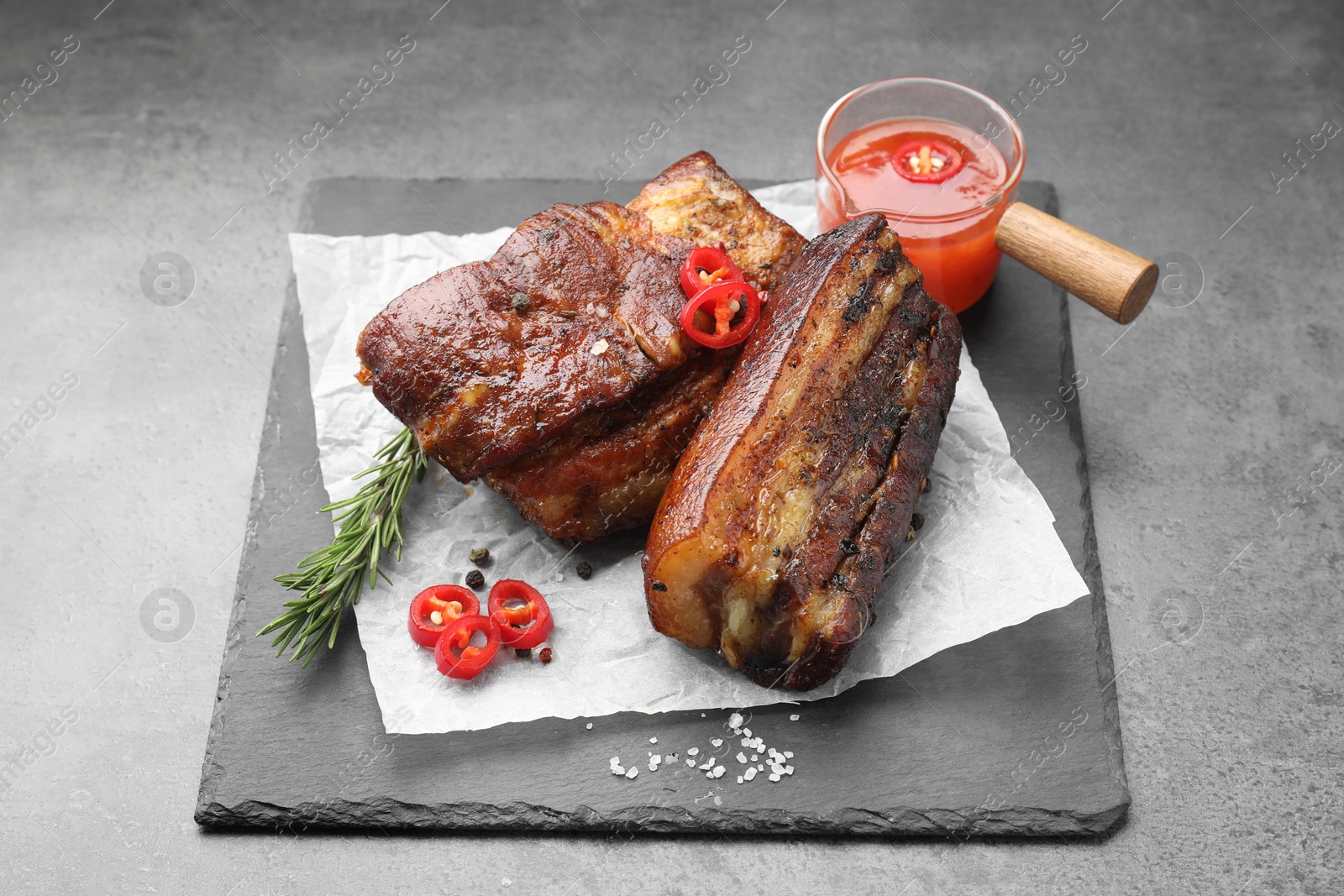 Photo of Pieces of baked pork belly served with sauce, rosemary and chili pepper on grey table