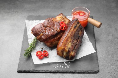 Photo of Pieces of baked pork belly served with sauce, rosemary and chili pepper on grey table