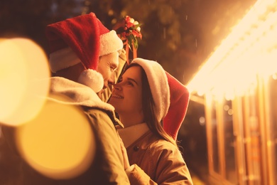 Happy couple in Santa hats standing under mistletoe bunch outdoors, bokeh effect