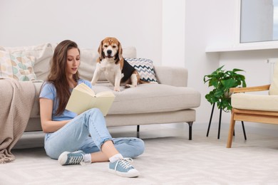 Beautiful young woman reading book near her cute Beagle dog on couch at home. Lovely pet