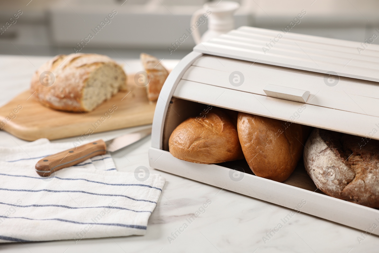 Photo of Wooden bread basket with freshly baked loaves and knife on white marble table in kitchen