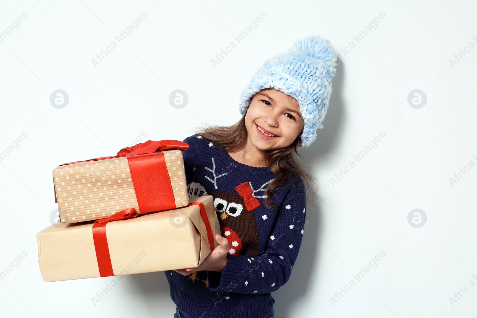 Photo of Cute little girl in Christmas sweater and knitted hat holding gifts on white background