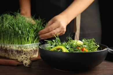 Photo of Woman picking fresh organic microgreen at wooden table, focus on bowl of delicious salad