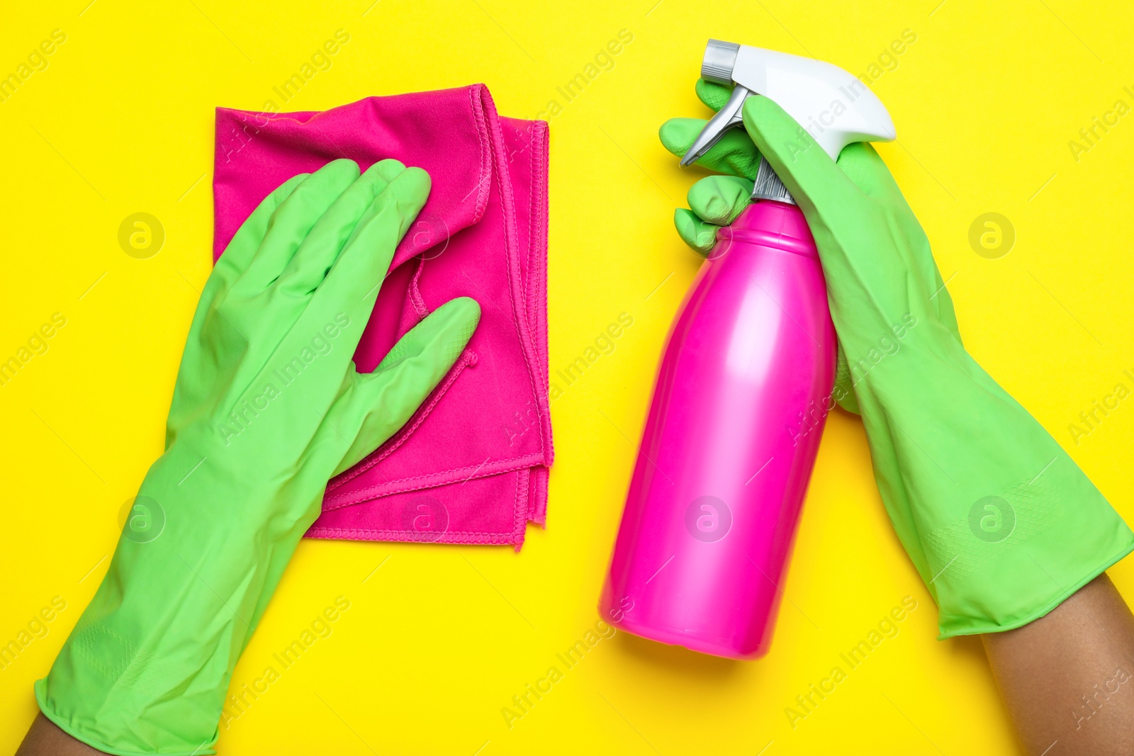 Photo of Woman in green rubber gloves with microfiber cloth and detergent on yellow background, top view