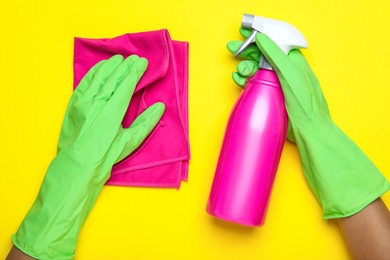 Woman in green rubber gloves with microfiber cloth and detergent on yellow background, top view