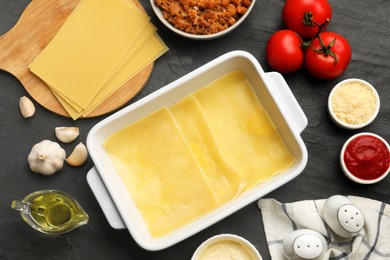 Photo of Cooking lasagna. Pasta sheets in baking tray and products on dark textured table, flat lay