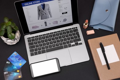Photo of Online store website on laptop screen. Computer, smartphone, credit cards, women's bag, stationery and lipstick on black background, above view