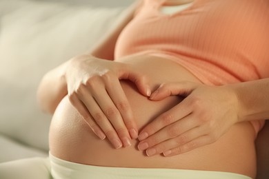 Photo of Pregnant woman making heart with her hands near belly indoors, closeup