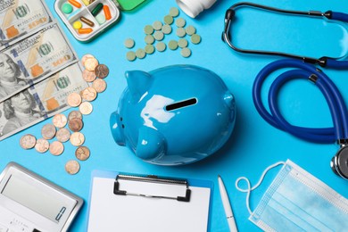 Ceramic piggy bank, stethoscope, money and pills on light blue background, flat lay. Medical insurance