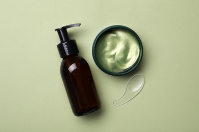 Photo of Under eye patches in jar with spatula and bottle of cosmetic product on olive background