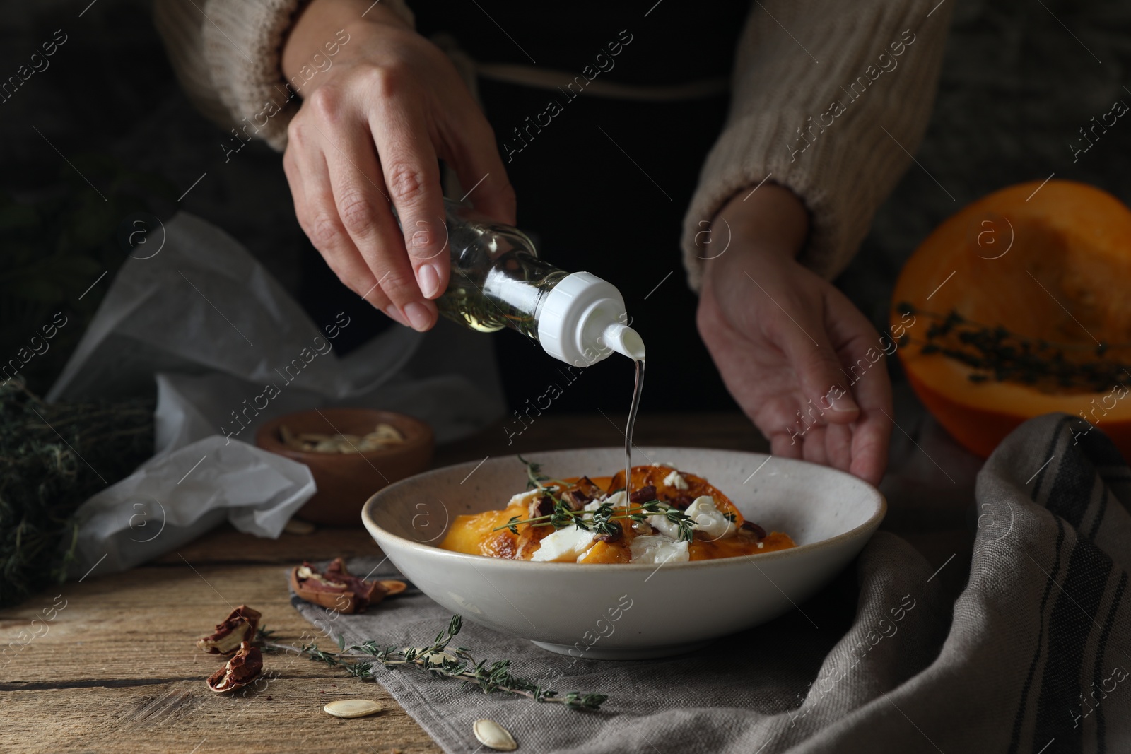 Photo of Woman adding cooking oil to freshly baked pumpkin slices with cheese, thyme and pecans at wooden table, closeup