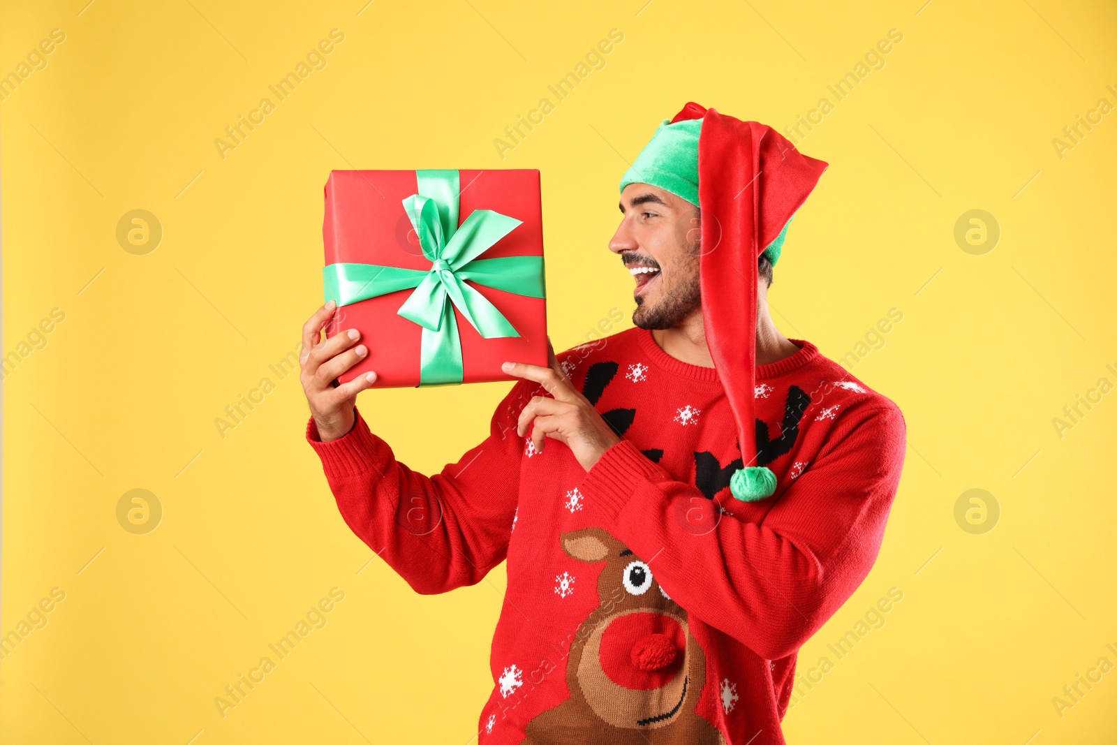 Photo of Happy man in Christmas sweater and elf hat holding gift box on yellow background