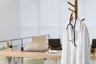 Photo of Modern doctor's workplace with wooden table and white coat hanging on rack in office