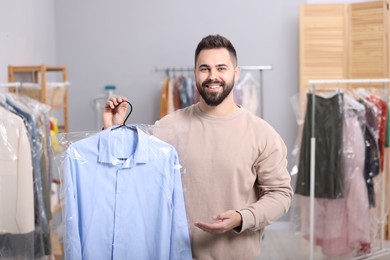 Photo of Dry-cleaning service. Happy man holding hanger with shirt in plastic bag indoors