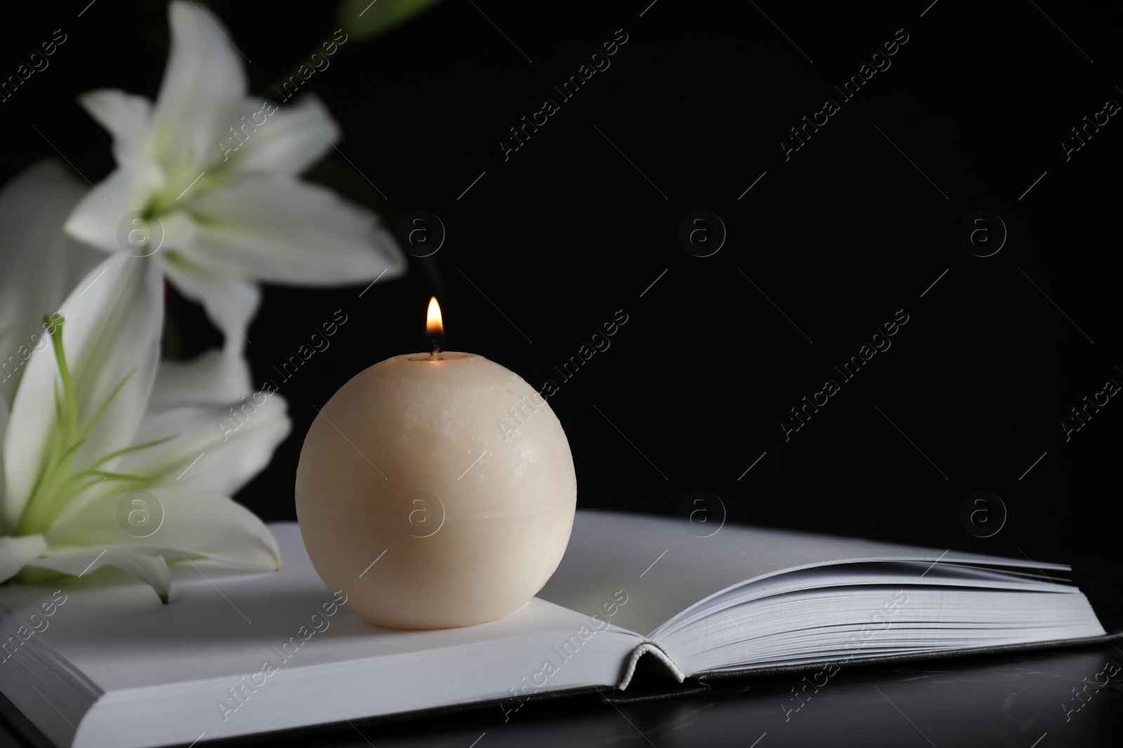 Photo of Burning candle, book and white lilies on table in darkness, closeup with space for text. Funeral symbol