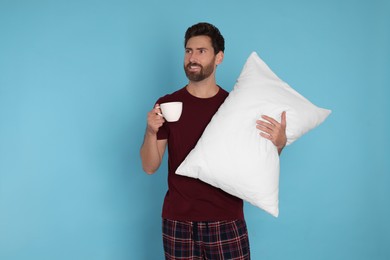 Handsome man with cup of hot drink and soft pillow on light blue background